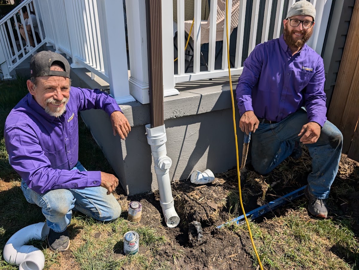 Two F.E. Johanssen and Sons crew members in branded purple shirts installing a downspout drain line beside a porch
